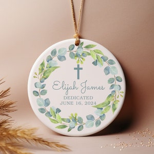May include: White ceramic ornament with a green eucalyptus wreath, a cross, and the text "Elijah James Dedicated June 16, 2024".