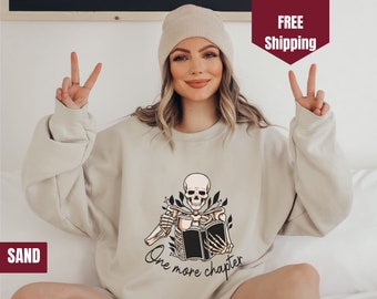 May include: A beige sweatshirt with a skeleton holding a book and a mug. The text on the sweatshirt reads "One more chapter".