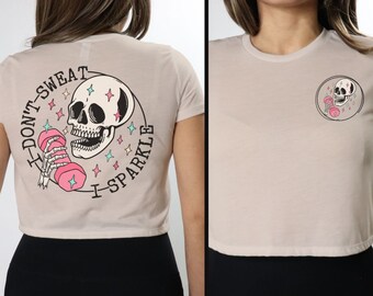 May include: A cropped, light beige t-shirt with a skull graphic and the text "I Don't Sweat I Sparkle" on the back. The front of the shirt has a smaller skull graphic with a dumbbell.