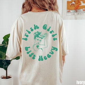 以下が含まれることがあります： クリーム色のTシャツに、緑色の女性がウェイトリフティングをしているグラフィックがプリントされています。グラフィックには、レトロなフォントで「Irish Girls Lift Heavy」と書いてあります。