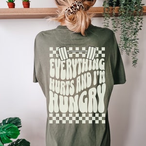 May include: Sage green t-shirt with a white graphic print of a barbell and the text "Everything Hurts and I'm Hungry". The graphic is in a retro style with a checkered background.
