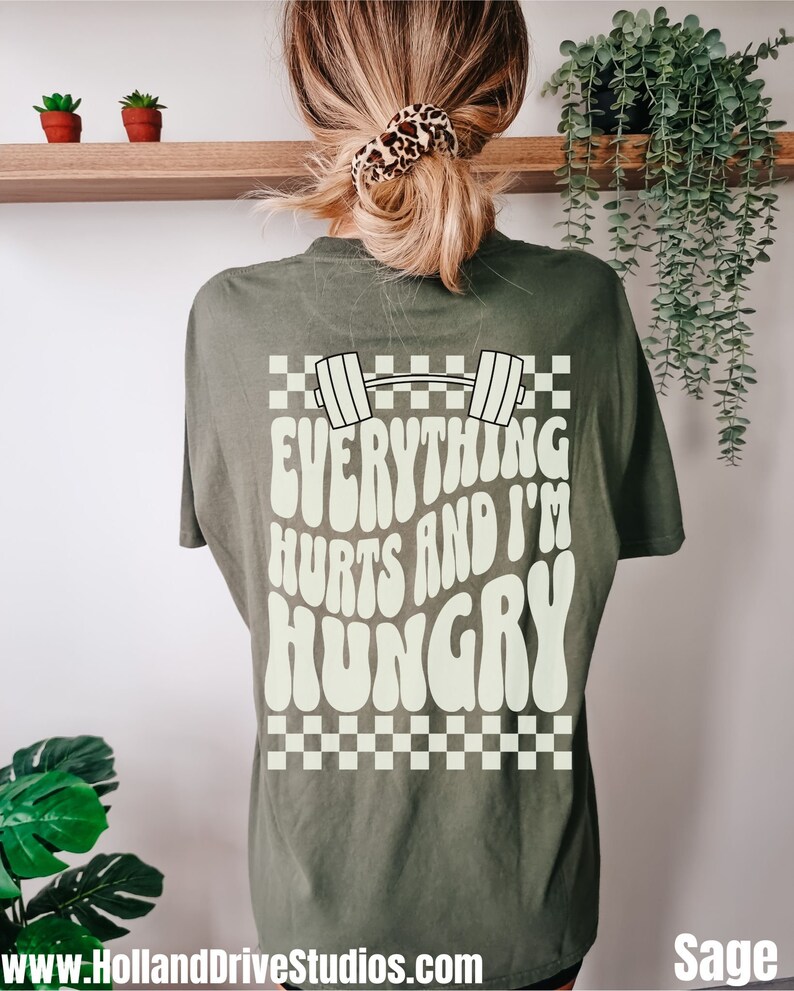 May include: Sage green t-shirt with a white graphic print of a barbell and the text "Everything Hurts and I'm Hungry". The graphic is in a retro style with a checkered background.