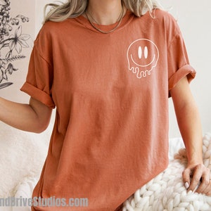 May include: A rust-colored t-shirt with a white dripping smiley face graphic.
