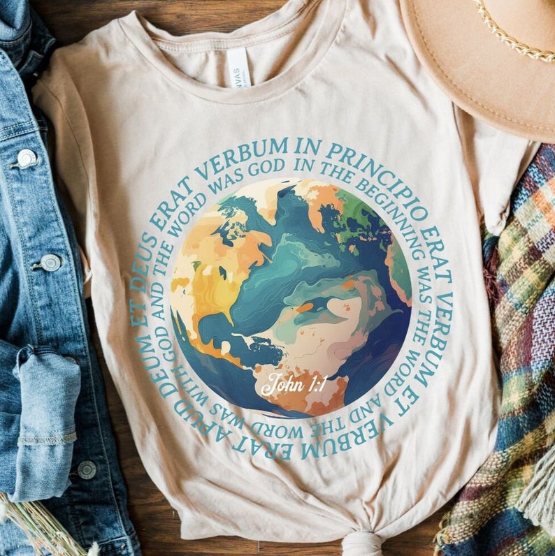 Op de afbeelding: Beige t-shirt met een kleurrijke illustratie van de aarde. Het ontwerp bevat een lichtblauwe cirkelvormige tekst met de woorden "VERBUM IN PRINCIPIO" en "John 1:1". Het shirt is gemaakt van een zacht materiaal.