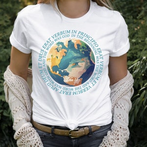 Op de afbeelding: Wit T-shirt met een kleurrijk wereldkaartontwerp met de tekst "VERBUM ERAT IN PRINCIPIO ERAT" en "John 1:1". Het ontwerp bevat tinten blauw, groen en geel. Het shirt is gemaakt van een zacht materiaal.