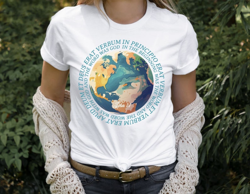 Op de afbeelding: Wit T-shirt met een kleurrijk wereldkaartontwerp met de tekst "VERBUM ERAT IN PRINCIPIO ERAT" en "John 1:1". Het ontwerp bevat tinten blauw, groen en geel. Het shirt is gemaakt van een zacht materiaal.