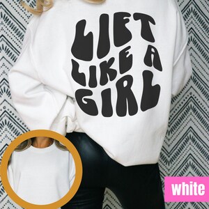 Puede incluir: Sudadera blanca con texto negro que dice "Lift Like A Girl" en la espalda. La sudadera se muestra en una modelo con pantalones negros.