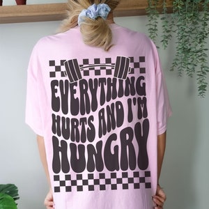 May include: Pink t-shirt with a black and white checkered pattern and the text "Everything Hurts and I'm Hungry" in a retro font. A black barbell is above the text.
