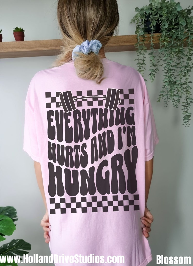 May include: Pink t-shirt with a black and white checkered pattern and the text "Everything Hurts and I'm Hungry" in a retro font. A black barbell is above the text.