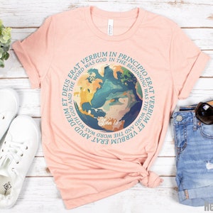 Op de afbeelding: Lichtperzikkleurig T-shirt met een cirkelvormige afbeelding van de aarde en een Latijnse quote. De tekst luidt: 'In het begin was het Woord, en het Woord was bij God.' Het shirt wordt gecombineerd met witte sneakers en een spijkerbroek.