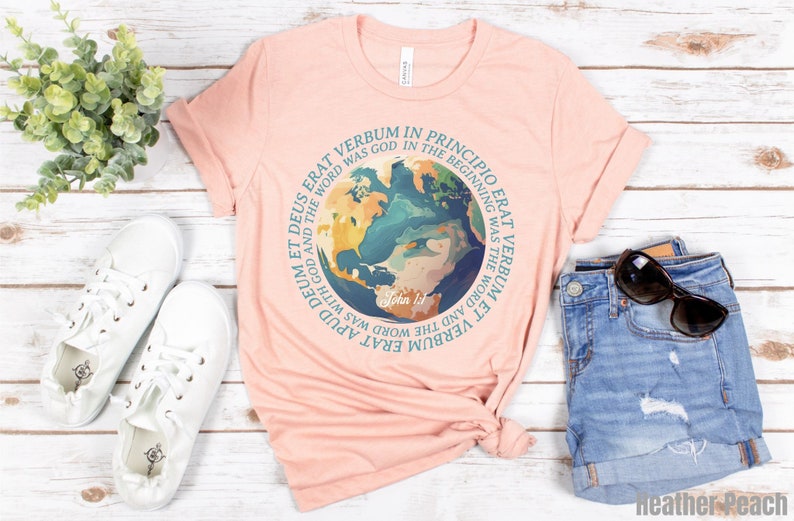 Op de afbeelding: Lichtperzikkleurig T-shirt met een cirkelvormige afbeelding van de aarde en een Latijnse quote. De tekst luidt: 'In het begin was het Woord, en het Woord was bij God.' Het shirt wordt gecombineerd met witte sneakers en een spijkerbroek.