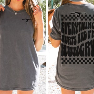 K&ouml;nnte beinhalten: Graues Oversize-T-Shirt mit einer Turner-Silhouette auf der Vorderseite und dem Text "Everything Hurts and I'm Hungry" auf der R&uuml;ckseite. Der Text ist in einer Retro-Schriftart mit einem karierten Muster.
