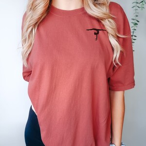 K&ouml;nnte beinhalten: Ein &uuml;berdimensioniertes T-Shirt in Rostfarbe mit einer schwarzen Silhouette einer Turnerin, die einen Handstand an den Stufenbarren macht.