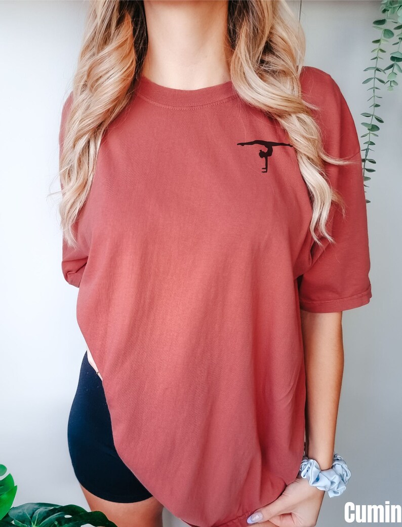 K&ouml;nnte beinhalten: Ein &uuml;berdimensioniertes T-Shirt in Rostfarbe mit einer schwarzen Silhouette einer Turnerin, die einen Handstand an den Stufenbarren macht.