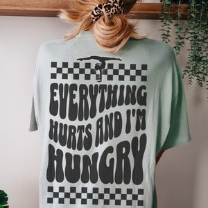 K&ouml;nnte beinhalten: Ein hellblaues T-Shirt mit einem schwarz-wei&szlig;en Karomuster oben und unten. Das Shirt hat eine schwarze Grafik einer Turnerin auf einem Schwebebalken mit dem Text "Everything Hurts and I'm Hungry" in einer Retro-Schrift.