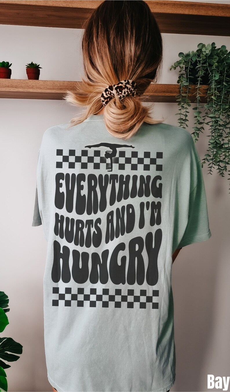 K&ouml;nnte beinhalten: Ein hellblaues T-Shirt mit einem schwarz-wei&szlig;en Karomuster oben und unten. Das Shirt hat eine schwarze Grafik einer Turnerin auf einem Schwebebalken mit dem Text "Everything Hurts and I'm Hungry" in einer Retro-Schrift.