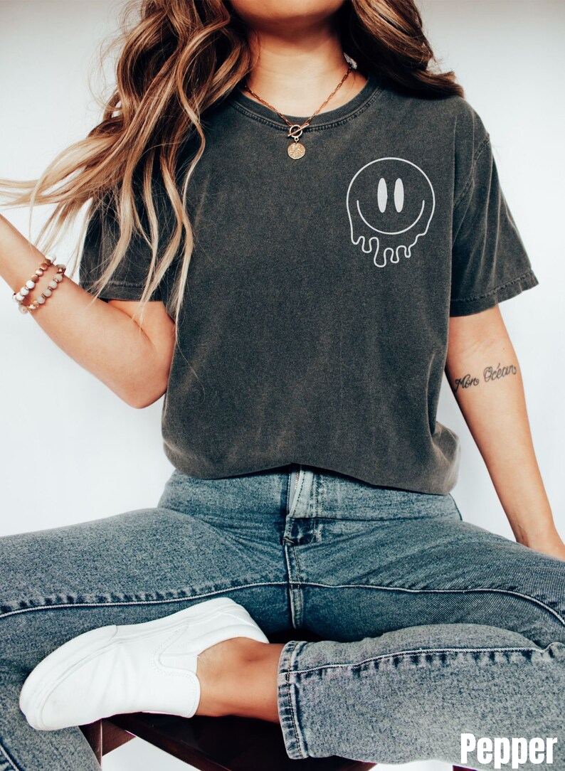 May include: A dark gray t-shirt with a white dripping smiley face graphic. The shirt is worn by a person with long blonde hair and a gold necklace. The person is wearing blue jeans and white sneakers.