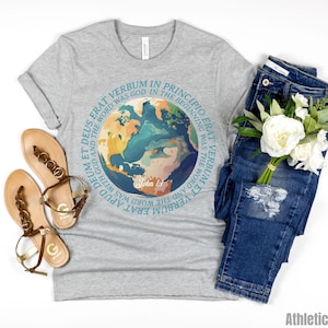Op de afbeelding: Een heather grijze t-shirt met een kleurrijk wereldbolontwerp en de tekst "IN THE BEGINNING WAS THE WORD" in een cirkelvormig patroon. De afbeelding bevat een blauwe jeans, sandalen en een boeket witte bloemen. De tekst "John 1:1" is ook zichtbaar.
