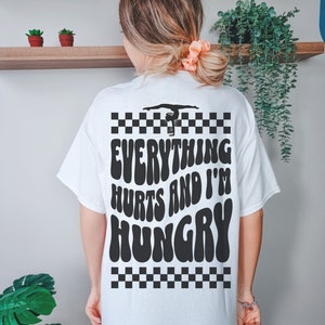 K&ouml;nnte beinhalten: Wei&szlig;es T-Shirt mit einem schwarz-wei&szlig;en Karomuster, einer Silhouette einer Turnerin auf einem Schwebebalken und dem Text "Everything Hurts and I'm Hungry" in einer Retro-Schrift.