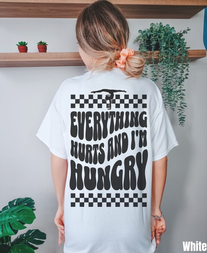 K&ouml;nnte beinhalten: Wei&szlig;es T-Shirt mit einem schwarz-wei&szlig;en Karomuster, einer Silhouette einer Turnerin auf einem Schwebebalken und dem Text "Everything Hurts and I'm Hungry" in einer Retro-Schrift.