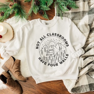 K&ouml;nnte beinhalten: Wei&szlig;er Pullover mit einem schwarz-wei&szlig;en Grafikdesign eines Wohnmobils in einer Berglandschaft. Der Text lautet "Not all classrooms have four walls".