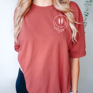 May include: A rust-colored t-shirt with a white dripping smiley face graphic.