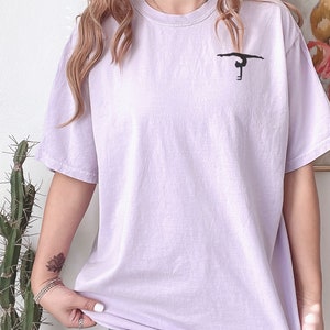 K&ouml;nnte beinhalten: Ein lavendelfarbenes T-Shirt mit einer schwarzen Silhouette einer Turnerin, die einen Handstand an den Stufenbarren macht.
