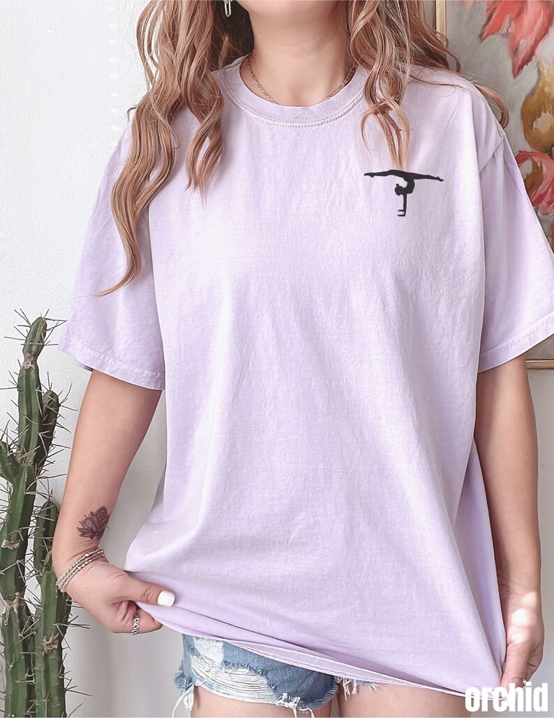 K&ouml;nnte beinhalten: Ein lavendelfarbenes T-Shirt mit einer schwarzen Silhouette einer Turnerin, die einen Handstand an den Stufenbarren macht.