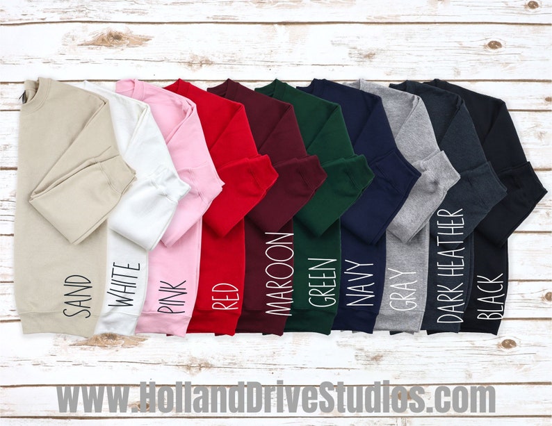 Puede incluir: Una fila de diez sudaderas con cuello redondo en varios colores, incluyendo arena, blanco, rosa, rojo, granate, verde, azul marino, gris, gris oscuro y negro. Las sudaderas est&aacute;n dispuestas sobre una superficie de madera blanca.