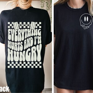 May include: Black t-shirt with a white graphic on the back that says "Everything Hurts and I'm Hungry" with a dumbbell and checkered pattern. The front of the shirt has a white dripping smiley face graphic.