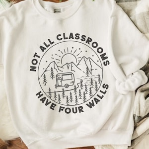 K&ouml;nnte beinhalten: Wei&szlig;es Sweatshirt mit einem schwarz-wei&szlig;en Grafikdesign eines Wohnmobils, das durch eine Berglandschaft f&auml;hrt. Der Text "Not all classrooms have four walls" ist in einem Kreis um die Grafik gedruckt.