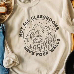 K&ouml;nnte beinhalten: Ein wei&szlig;er Pullover mit einem schwarzen Grafikdesign eines Wohnmobils, das durch eine Berglandschaft f&auml;hrt. Die Grafik ist von einem Kreis mit dem Text "Not all classrooms have four walls" umgeben.