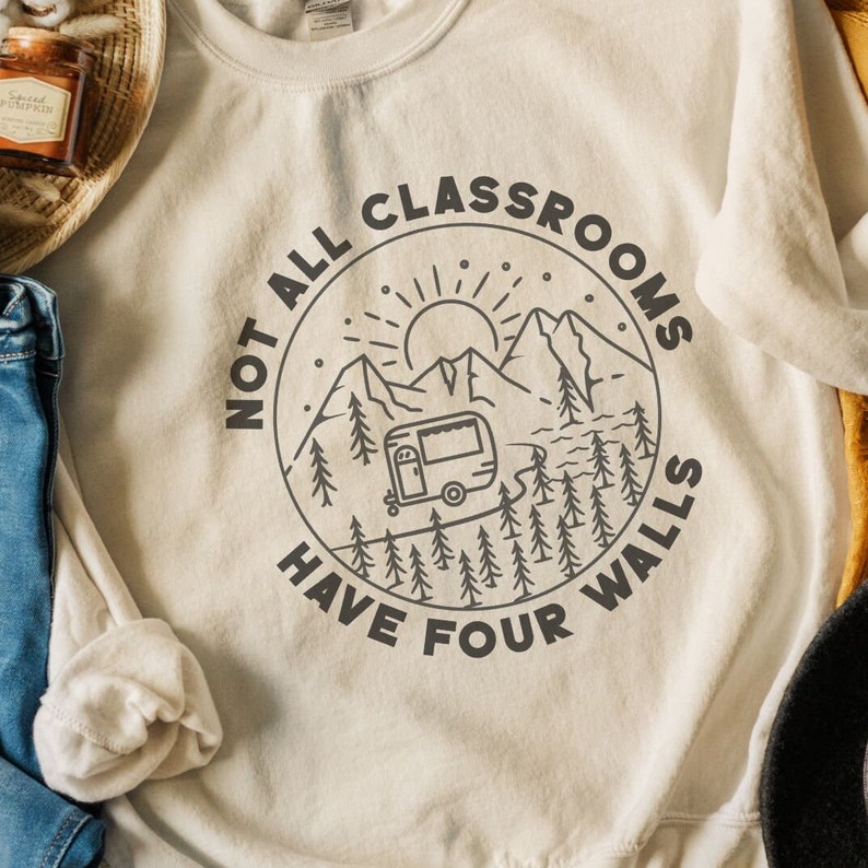 K&ouml;nnte beinhalten: Ein wei&szlig;er Pullover mit einem schwarzen Grafikdesign eines Wohnmobils, das durch eine Berglandschaft f&auml;hrt. Die Grafik ist von einem Kreis mit dem Text "Not all classrooms have four walls" umgeben.