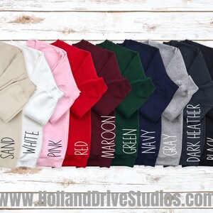 May include: A row of ten crewneck sweatshirts in various colours, including sand, white, pink, red, maroon, green, navy, grey, dark heather, and black. The sweatshirts are laid out on a white wooden surface.