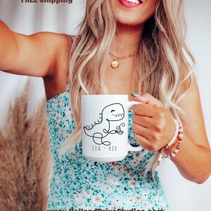 Könnte beinhalten: Weiße Keramiktasse mit einer schwarzen Strichzeichnung eines Dinosauriers, der eine Teetasse hält, mit dem Text "TEA REX". Die Tasse hat einen großen Henkel. Ein lustiger und skurriler Artikel.