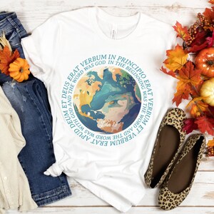Op de afbeelding: Wit T-shirt met een wereldbol-afbeelding en Latijnse tekst, waaronder "In het begin was het Woord." Het shirt wordt gecombineerd met een blauwe jeans, een cr&egrave;mekleurig vest en luipaardprint ballerina's. Herfstbladeren en pompoenen vullen de sc&egrave;ne aan.