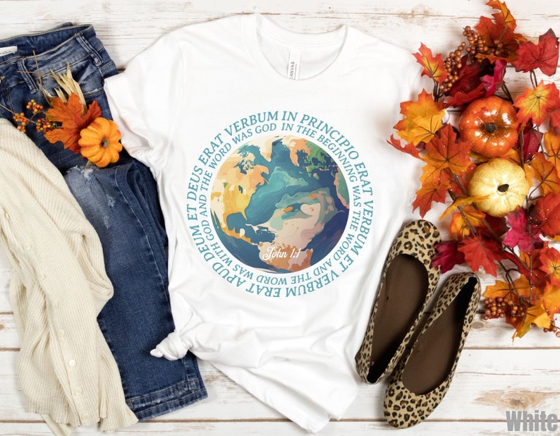 Op de afbeelding: Wit T-shirt met een wereldbol-afbeelding en Latijnse tekst, waaronder "In het begin was het Woord." Het shirt wordt gecombineerd met een blauwe jeans, een cr&egrave;mekleurig vest en luipaardprint ballerina's. Herfstbladeren en pompoenen vullen de sc&egrave;ne aan.