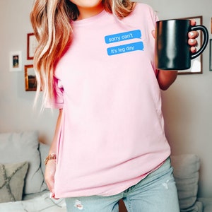 Może przedstawiać: R&oacute;żowy t-shirt z niebieskim graficznym elementem w kształcie dymku z tekstem "sorry can't it's leg day".