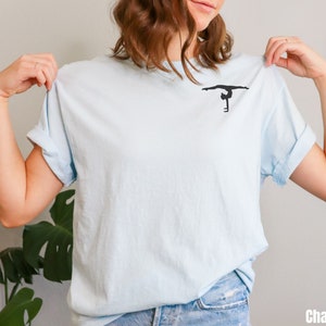 K&ouml;nnte beinhalten: Ein hellblaues T-Shirt mit einer schwarzen Silhouette einer Turnerin, die einen Handstand an den Stufenbarren macht.