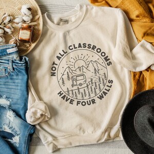 K&ouml;nnte beinhalten: Ein beiger Pullover mit einem grafischen Design eines Wohnmobils in einer Berglandschaft. Der Text lautet "Not all classrooms have four walls".