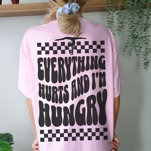 K&ouml;nnte beinhalten: Rosa T-Shirt mit einer schwarzen Silhouette einer Turnerin auf einem Schwebebalken und dem Text "Everything hurts and I'm hungry" in einer Retro-Schrift.