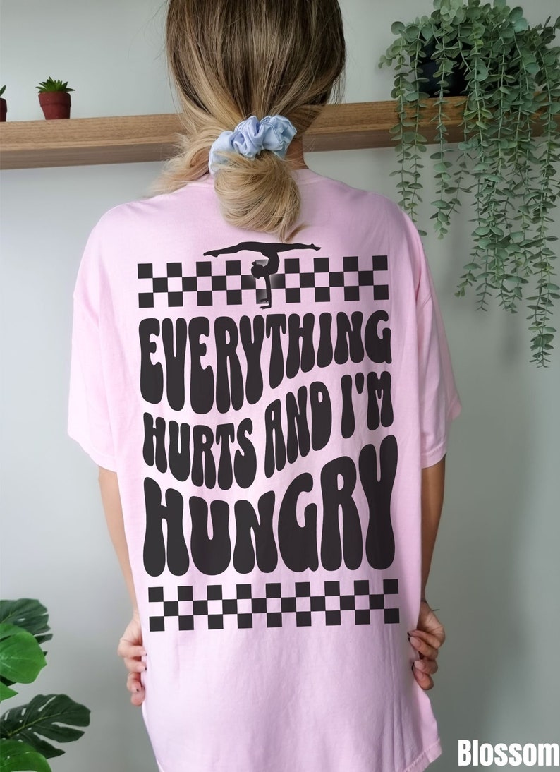 K&ouml;nnte beinhalten: Rosa T-Shirt mit einer schwarzen Silhouette einer Turnerin auf einem Schwebebalken und dem Text "Everything hurts and I'm hungry" in einer Retro-Schrift.
