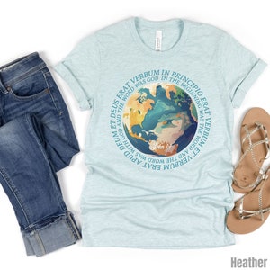 Op de afbeelding: Een lichtblauw t-shirt met een cirkelvormige afbeelding van de aarde en de tekst "In het begin was het Woord". Het shirt wordt gecombineerd met een blauwe jeans en gouden sandalen. Het shirt is Heather Ice Blue.