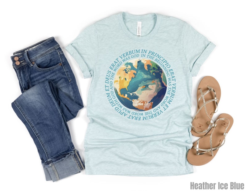 Op de afbeelding: Een lichtblauw t-shirt met een cirkelvormige afbeelding van de aarde en de tekst "In het begin was het Woord". Het shirt wordt gecombineerd met een blauwe jeans en gouden sandalen. Het shirt is Heather Ice Blue.