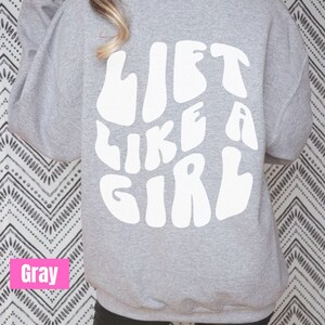 Puede incluir: Sudadera gris con el texto "LIFT LIKE A GIRL" en blanco, con letras de estilo retro.