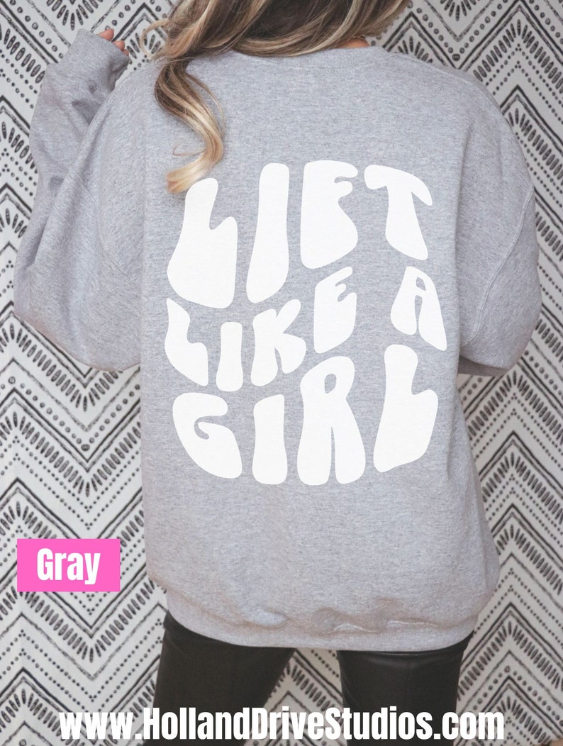 Puede incluir: Sudadera gris con el texto "LIFT LIKE A GIRL" en blanco, con letras de estilo retro.