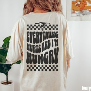 K&ouml;nnte beinhalten: Ein wei&szlig;es T-Shirt mit einer schwarzen Grafik, auf der "Alles tut weh und ich habe Hunger" steht, mit einer Silhouette einer Turnerin auf einem Schwebebalken &uuml;ber dem Text. Die Grafik ist von einem schwarz-wei&szlig;en Karomuster umgeben.
