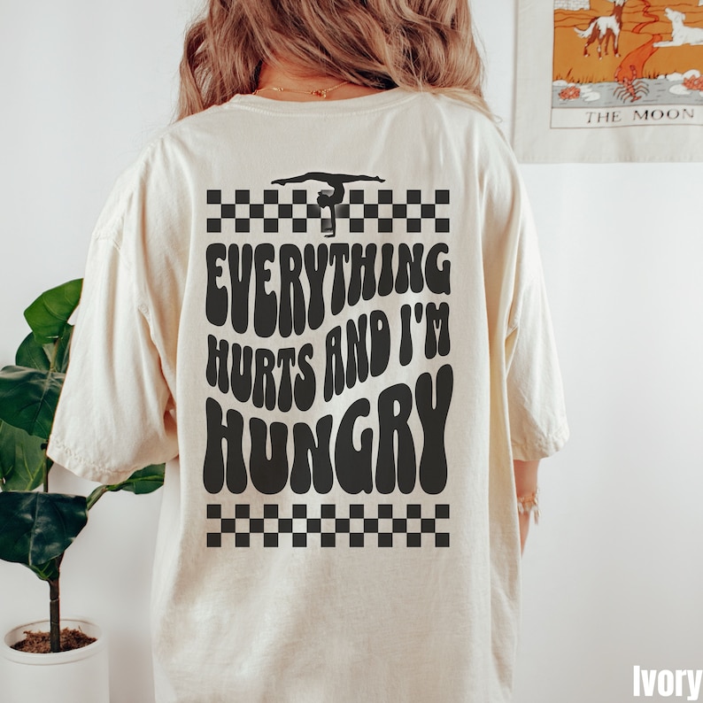 K&ouml;nnte beinhalten: Ein wei&szlig;es T-Shirt mit einer schwarzen Grafik, auf der "Alles tut weh und ich habe Hunger" steht, mit einer Silhouette einer Turnerin auf einem Schwebebalken &uuml;ber dem Text. Die Grafik ist von einem schwarz-wei&szlig;en Karomuster umgeben.