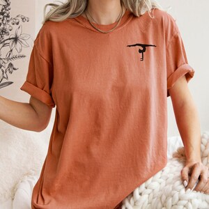 K&ouml;nnte beinhalten: Ein rostfarbenes T-Shirt mit einer schwarzen Silhouette einer Turnerin auf einem Schwebebalken.