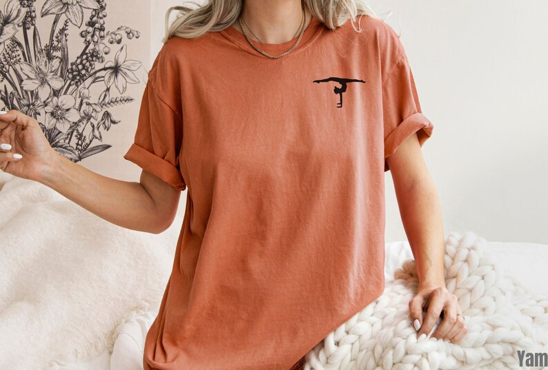 K&ouml;nnte beinhalten: Ein rostfarbenes T-Shirt mit einer schwarzen Silhouette einer Turnerin auf einem Schwebebalken.